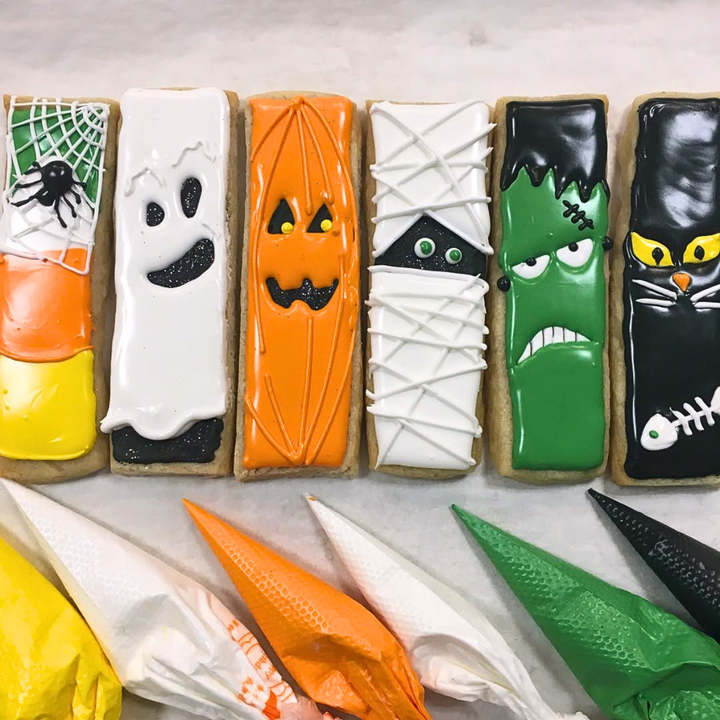 Six Halloween-themed rectangular sugar cookies decorated with colorful icing in candy corn, ghost, pumpkin, mummy, Frankenstein, and cat designs, arranged on a light marble surface with piping bags below.