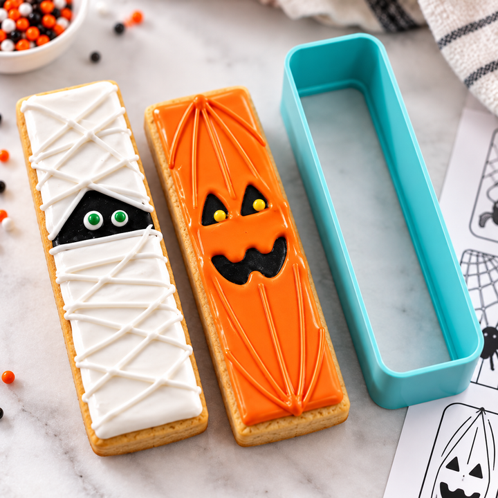 A rectangular turquoise 3d printed cookie cutter shown beside two decorated Halloween cookies on a marble surface. One cookie is white iced like a mummy with black eyes, and the other is orange iced like a jack-o&